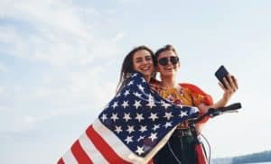 Two patriotic cheerful women with bike and USA flag in hands makes selfie